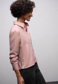 Blusa rosa chiaro a maniche lunghe con bottoni frontali, dettaglio a strisce laterali e silhouette rilassata. La texture liscia si abbina a uno stile informale.
