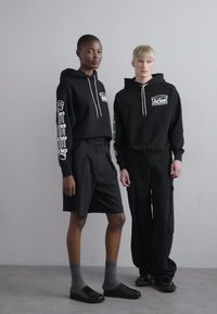 Twee modellen dragen zwarte hoodies met witte graphics, de ene gecombineerd met zwarte shorts en de andere met cargobroeken. Beide hebben donkere schoenen.