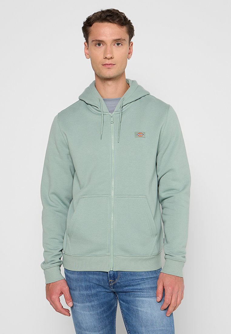 Sudadera con cremallera de color verde claro, fabricada en material suave, con un bolsillo tipo canguro, capucha ajustable con cordón y pequeño logotipo en el pecho.