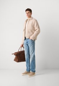 Chaqueta de mezcla de lana beige con dos bolsillos frontales, camisa crema, jeans azul claro, zapatos de ante marrón claro y bolsa de viaje de cuero marrón.