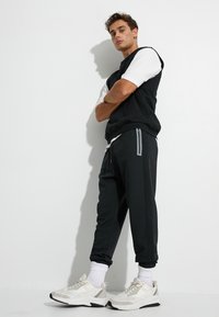 Sort ærmeløs sweatshirt over en hvid t-shirt, parret med sorte joggers med grå striber, og lyse sneakers med en tekstureret finish.