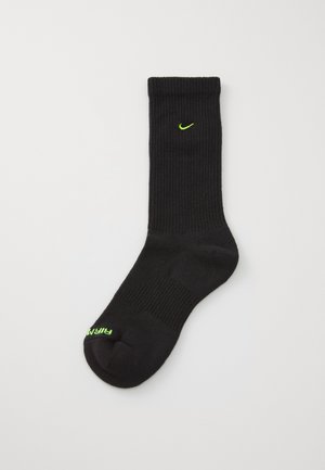 Calcetín negro de canalé con un logo de Nike en verde neón en la parte superior y la palabra "FIRM" en verde en la zona de los dedos. Hecho de material suave y elástico.