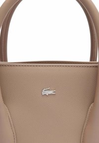 Sac à main beige avec une finition texturée, poignées supérieures arrondies, et un logo crocodile argenté à l'avant. Présente un design épuré et des accents lisses.