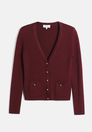 Femme portant une robe bordeaux et un cardigan avec un chemisier à nœud, tenant un sac à main noir et portant des bottes noires montant jusqu'aux genoux.