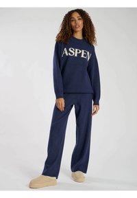 Felpa lavorata a maglia di colore blu navy con la scritta "ASPEN" in crema, abbinata a pantaloni larghi nella stessa tonalità. Modello con scarpe beige senza lacci.