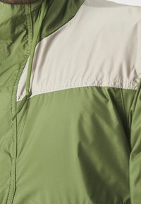 Chaqueta verde con un panel superior de color beige claro, tela ligera, capucha ajustable con cordón y textura suave.