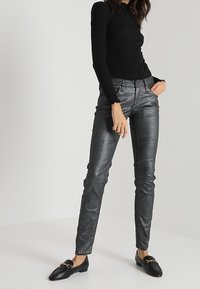 Pantalons skinny gris métallique avec une texture lisse, présentant une taille mi-haute, des passants de ceinture et cinq poches, associés à des ballerines noires.