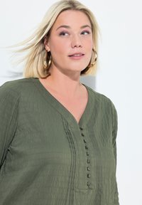 Olivgrüne, strukturierte Bluse mit V-Ausschnitt, vertikalen Falten und sieben Frontknöpfen. Weiches Gewebe mit einem leichten Erscheinungsbild.