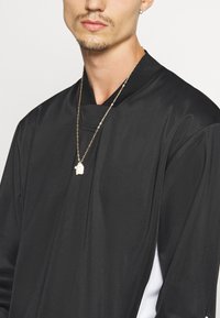 Haut noir à manches longues avec un col montant et des accents blancs. Porté avec une chaîne en or ornée d'un pendentif. Texture lisse.