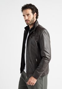 Bruine leren jas met een ritsopening aan de voorkant, hoge kraag en ritszakken; gestructureerde afwerking met een aansluitende pasvorm en schouderdetails.