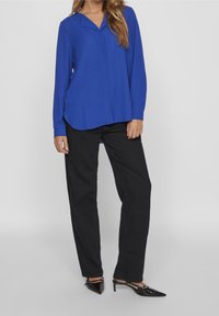 Blouse bleu roi à manches longues avec un col en V et une fermeture à boutons sur le devant, associée à un pantalon noir fuselé et des escarpins noirs à bouts pointus.