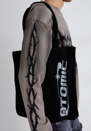 Juodas drobės rankinis krepšys su blizgiu sidabriniu "Atomic" logotipu, turi struktūrizuotą formą ir plačius hermetinius rankenėles; tekstūruotas pilkas megztinis su geometriniu raštu.