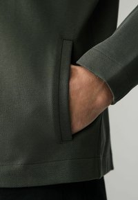 Tejido verde texturizado con patrón diagonal, que presenta un bolsillo lateral. La manga tiene un diseño ligeramente entallado. Fondo liso y oscuro.