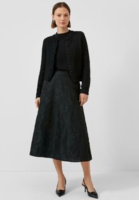 Schwarze gestrickte Cardigan mit rundem Ausschnitt und Knopfleiste, kombiniert mit einem schwarzen Midirock mit Textur und floralem Muster. Schwarze Absätze.