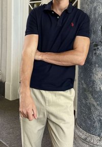 Jeune homme portant un polo bleu marine foncé et un pantalon beige clair, debout à l'intérieur près d'une colonne en marbre gris et un cadre de porte blanc.