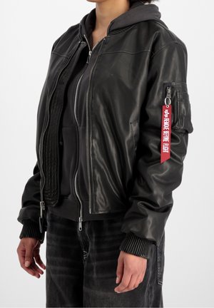 Person trägt eine schwarze Leder-Bomberjacke mit rotem "Remove Before Flight" Anhänger am Ärmel und einen schwarzen Zip-Hoodie darunter.