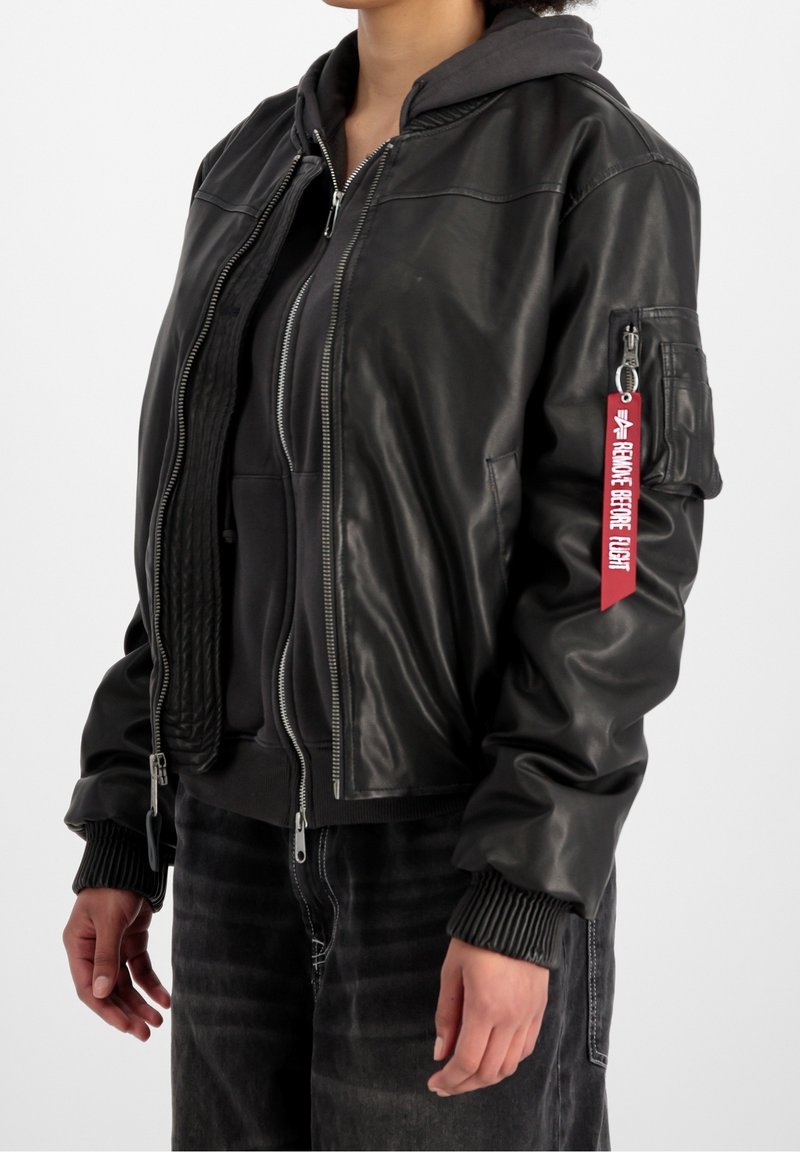 Personne portant une veste bomber en cuir noire avec une étiquette rouge "Remove Before Flight" sur la manche et un sweat à capuche noir zippé en dessous.