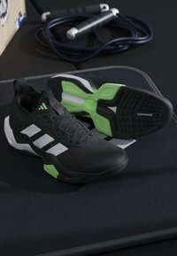 Paire de chaussures de sport noires avec des rayures blanches et des semelles vertes, posée sur un tapis noir avec une corde à sauter en arrière-plan.