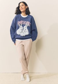 Marineblauwe sweatshirt met konijnenafbeelding en tekst "ONGELOOFLIJK", gecombineerd met lichtroze joggerbroek. Model staat nonchalant met handen in de zakken.