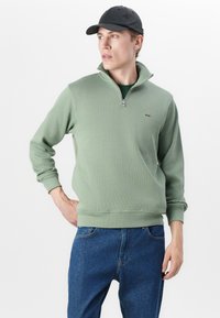 Groene geribbelde sweatshirts met een kwart ritssluiting, gemaakt van zacht materiaal, met een hoge kraag en geborduurd logo. Gepaard met blauwe spijkerbroeken.