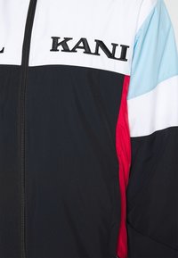 Väriblokki tuulitakki mustassa, valkoisessa, vaaleansinisessä ja punaisessa. Edessä vetoketju ja rintapielessä musta kirjailtu "KANI"-logo. Sileä pinta.