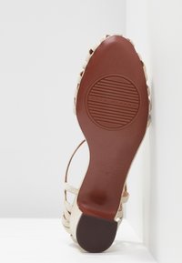 Beige sandál s tkaným textilním svrškem, otevřenou špičkou a hnědou koženou podrážkou se zakulaceným logem. Ploch�ý podpatek a žebrovaná textura.