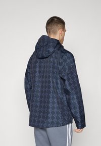 Chaqueta de agua en color marino con un diseño de logo estampado, que cuenta con capucha y mangas largas. La parte trasera presenta un corte relajado y un dobladillo curvado.