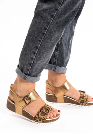 Braune und goldene Keilsandalen mit Leopardenmuster, Schnallendetail und strukturiertem Korksohle. Getragen mit hochgekrempelten grauen Jeans.