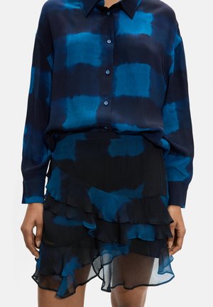 Kobieta w niebiesko-czarnej bluzce na guziki oraz pasującej warstwowej spódnicy z falbankami wykonanej z prześwitującego materiału.