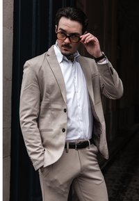 Mann i beige dress og hvit skjorte som justerer brune solbriller, stående mot en mørk urban bakgrunn med hånden i lomma.