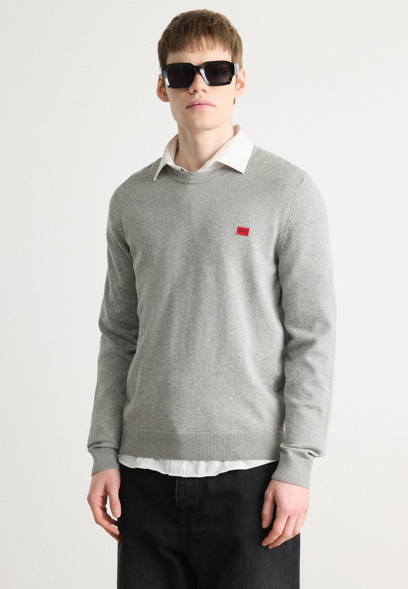 Siv sweater z okroglim izrezom in rebrastimi manšetami, z majhno rdečo logotipno nalepko. Obut nad belim srajčnim ovratnikom in črnimi džinsom.