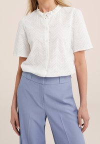 Vit kortärmad blus med zigzagmönster, med volangkrage och knappframslutning. I kombination med ljusblå skräddarsydda byxor.