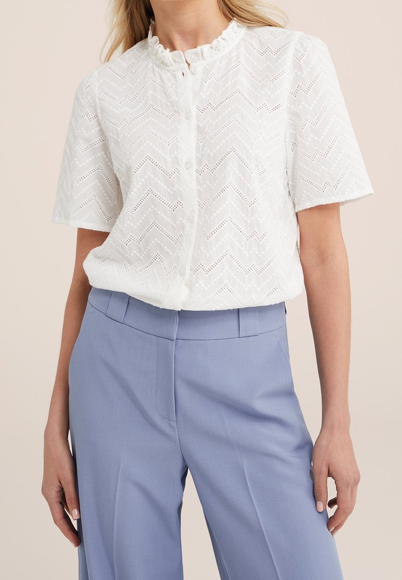 Vit kortärmad blus med zigzagmönster, med volangkrage och knappframslutning. I kombination med ljusblå skräddarsydda byxor.