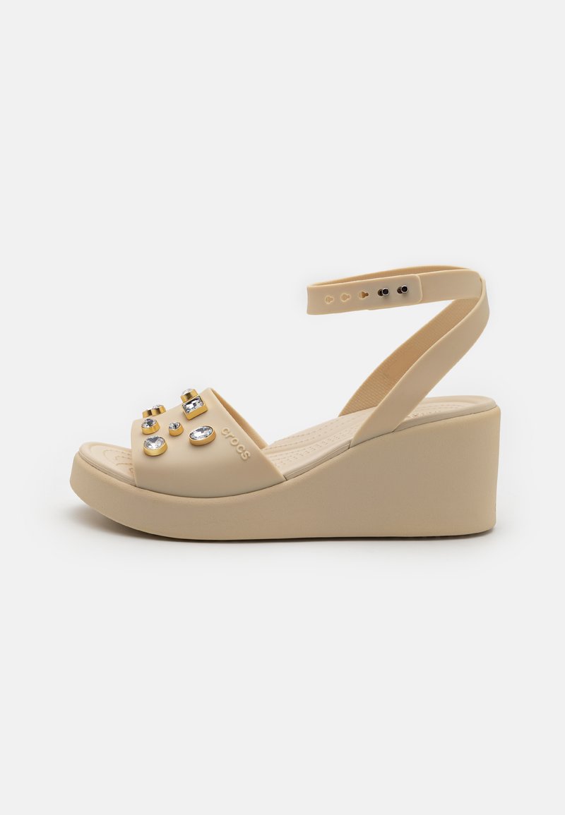 Sandale compensée beige avec des accents de strass, dotée d'une large sangle sur le bout du pied et d'une sangle de cheville ajustable pour un bon maintien. Texture douce, design léger.
