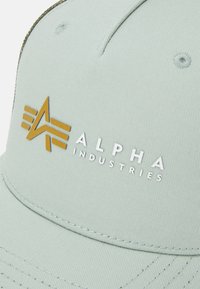 Lys grønn caps med teksturert stoff. Har en metallisk gulllogo og hvit tekst "ALPHA INDUSTRIES" på forsiden.