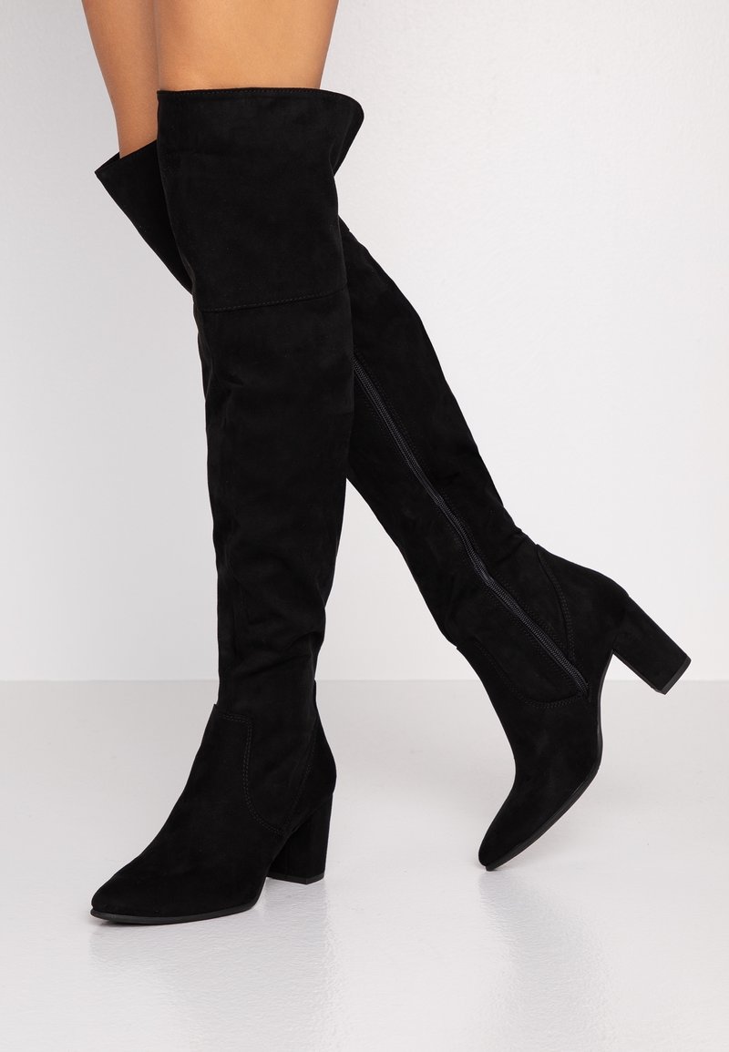 Jalkoja, joissa on mustat mokkanahkaiset polven ylle ulottuvat saappaat, joissa on leveät korot ja sivuvetoketjut, aseteltuna yksinkertaista vaaleaa taustaa vasten.