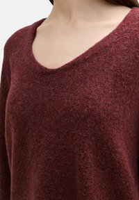 Bordeaux gebreide trui met een brede V-hals, zachte textuur en een losse fit, die de warmte en gezellige uitstraling van de stof benadrukt.