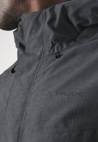 Gray jacket with a high collar, featuring a textured fabric, front zip, and adjustable closures. Logo visible on the left side.