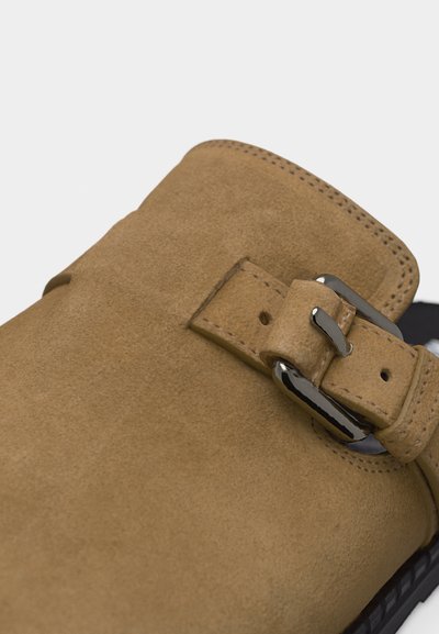 Gros plan d'une chaussure en daim beige avec un détail de sangle à boucle métallique et des coutures visibles sur la tige et le bord.
