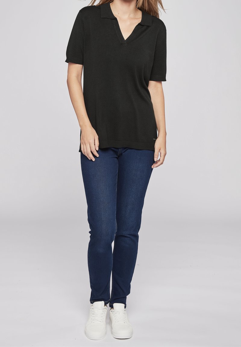 Svart kortärmad polo skjorta med v-ringning, tillsammans med mörkblå jeans och vita sneakers, visad mot en neutral bakgrund.