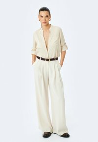 Kremowa koszula z guzikami, z podwiniętymi rękawami, w połączeniu z beżowymi spodniami o wysokim stanie i szerokich nogawkach oraz brązowym paskiem. Czarne buty.