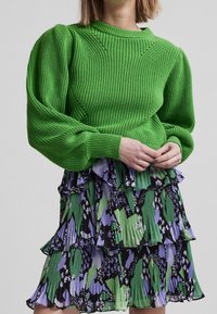 Pull vert en tricot avec manches bouffantes, associé à une robe à volants avec un motif floral en noir, vert et violet.