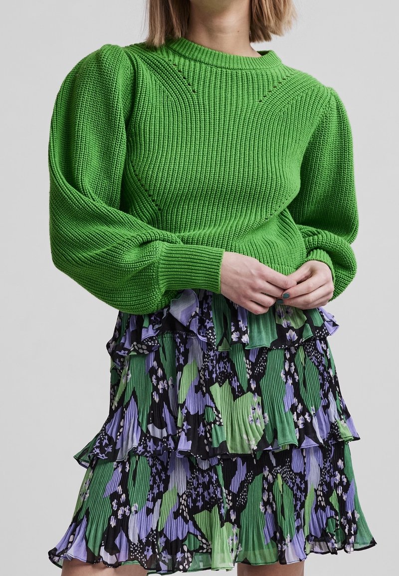 Pull vert en tricot avec manches bouffantes, associé à une robe à volants avec un motif floral en noir, vert et violet.