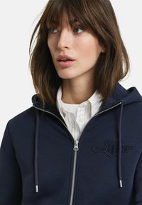 Nuori nainen, jolla on ruskeat hiukset, pukeutuneena tummansiniseen vetoketjulliseen huppariin valkoisen kauluspaitaisen paidan päällä, katse hieman sivulle yksiväristä taustaa vasten.