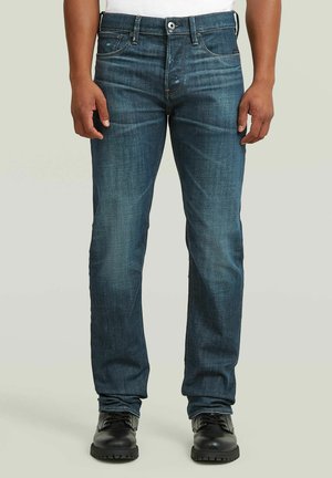 Mann trägt eng anliegende, dunkelblaue Jeans mit dezentem Verblassen und schwarze Lederstiefel, steht vor einem schlichten hellen Hintergrund.