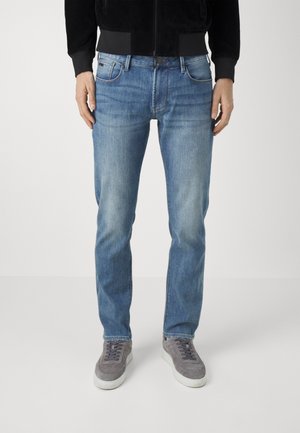 Mężczyzna ubrany w jasnoniebieskie, proste dżinsy, czarną kurtkę i szare trampki, stojący na jednolitym białym tle.