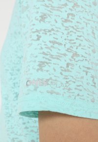 Ljus turkos t-shirt med ett subtilt texturerat mönster, korta ärmar och en etikett som visar "ONLY PLAY" i silver vid fållen.