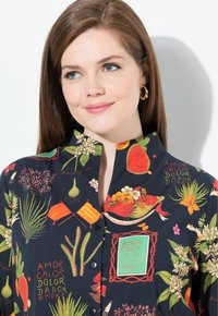 Camicia a collo con bottoni in tessuto scuro, caratterizzata da vivaci stampe botaniche e testo, con colori che includono verde, arancione, giallo e rosso.