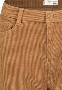Braune Cordhosen mit vertikaler Rippstruktur, vorderen Taschen und einem Knopfverschluss, versehen mit einem Etikett mit der Aufschrift "EIGHT2NINE SMALL."