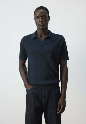 Homme à la peau foncée portant un polo en maille bleu foncé à manches courtes et un jean foncé, debout devant un fond gris clair.